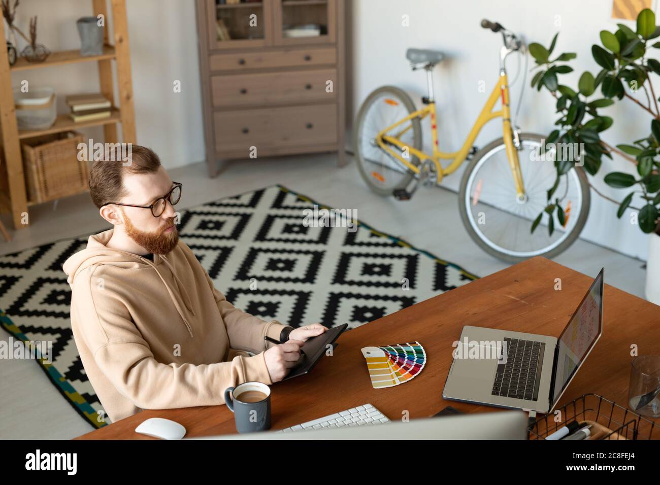 Ilustrador ocupado en hoodie sentado en la oficina en casa y haciendo boceto usando la tableta de dibujo Foto de stock