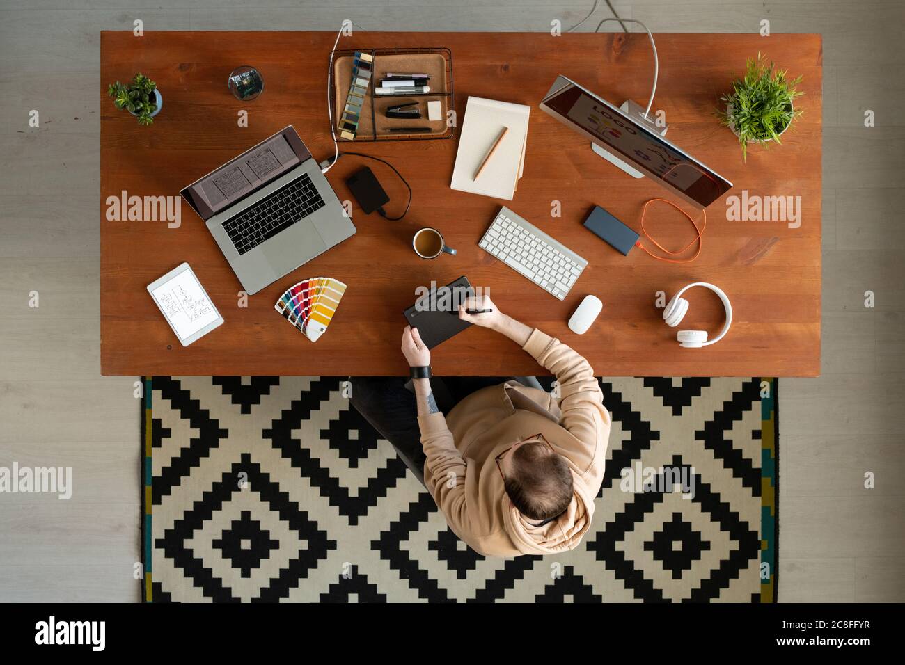 Vista superior de la interfaz de diseñador en hoodie sentado en el escritorio de madera lleno de dispositivos y dibujo de croquis en la tableta del digitalizador Foto de stock