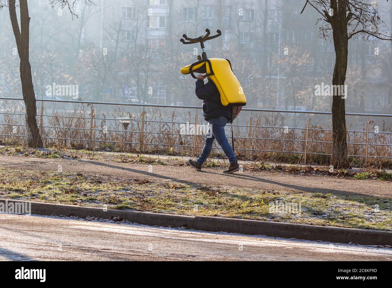 Temprano por la mañana, un joven con una mochila lleva una silla de oficina grande sobre su cabeza. Sillón amarillo brillante en la niebla de invierno. Foto de stock