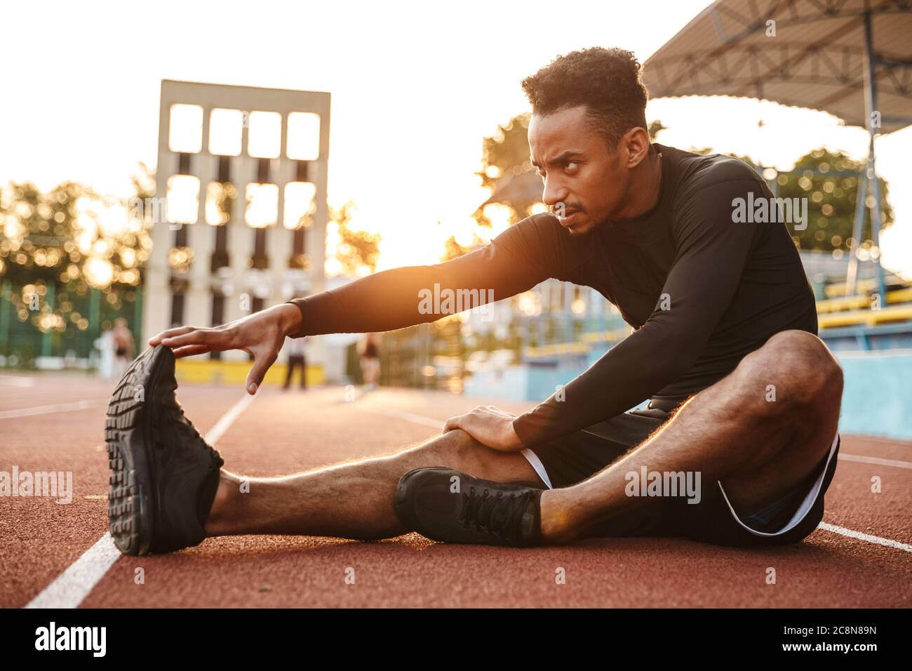 Imagen de fuerte hombre afroamericano estirando su cuerpo mientras está sentado en la cancha de deportes al aire libre Foto de stock