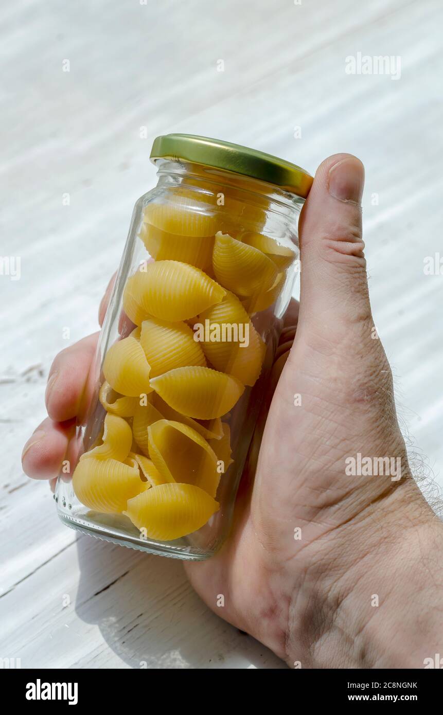 La mano macho sostiene un tarro de vidrio seco de Conchiglie. Porciones de pasta cruda en la mano de un adulto caucásico masculino. Embalaje ecológico para almacenar foo Foto de stock