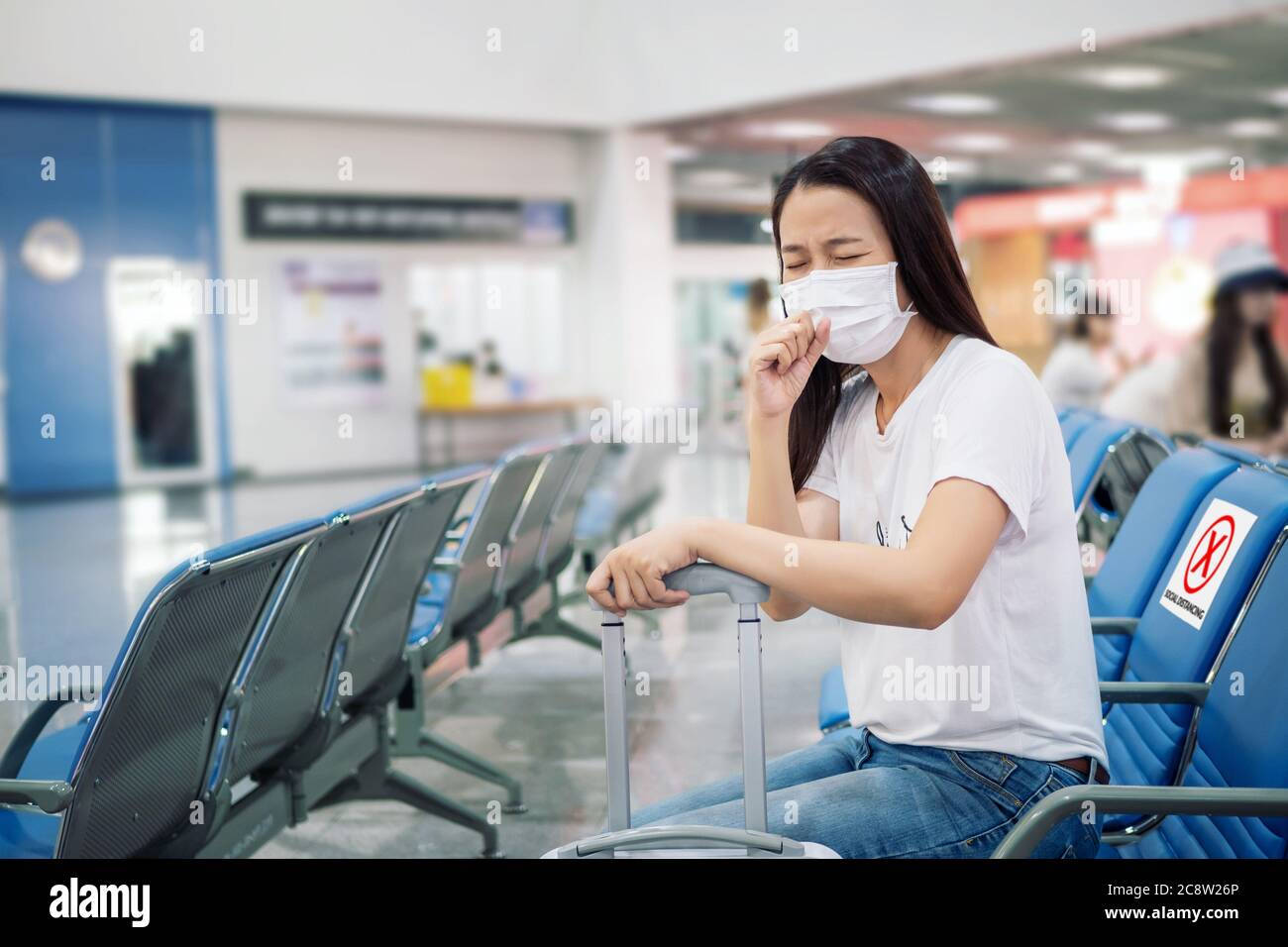 Turista asiático sentirse enfermo, toser, usar máscara y sentarse en silla con distanciamiento social para prevenir una pandemia durante los viajes en la terminal del aeropuerto. Foto de stock