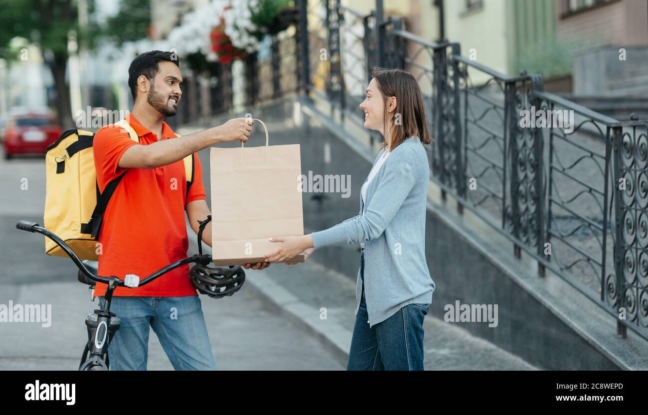 Excelente entrega al cliente en la ciudad. Un hombre sonriente con mochila y bicicleta le da una bolsa de papel a una chica feliz Foto de stock