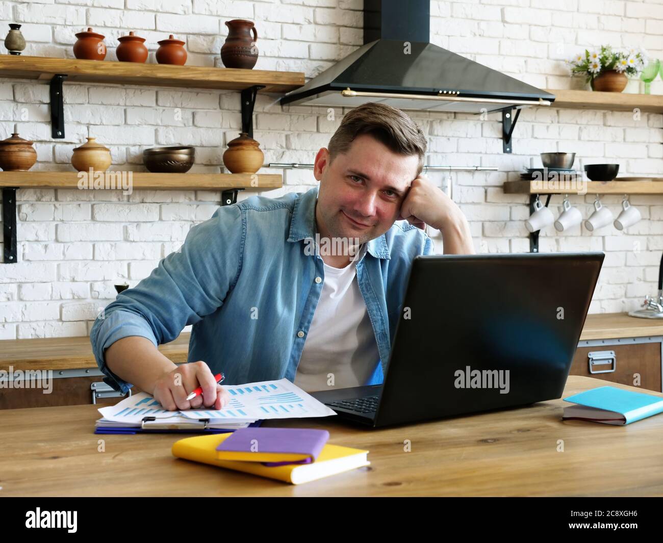 El freelancer trabaja remotamente desde casa. Un hombre con un ordenador portátil en la cocina. Foto de stock