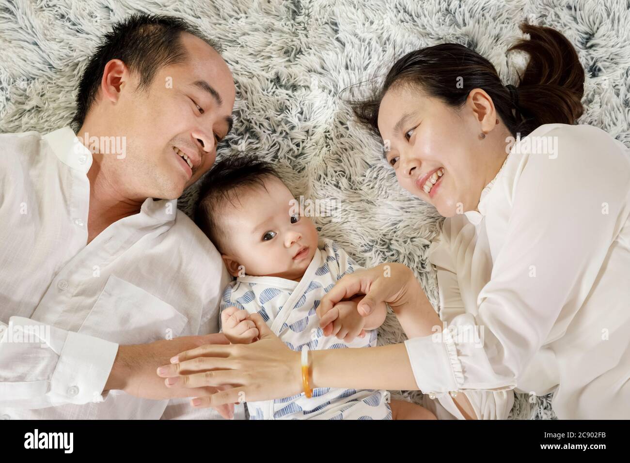Padres asiáticos jugando con el concepto de bebé, joven vietnamita hombre y mujer sonriendo e interactuando con un niño que se pone sobre una alfombra blanca, familia juntos Foto de stock
