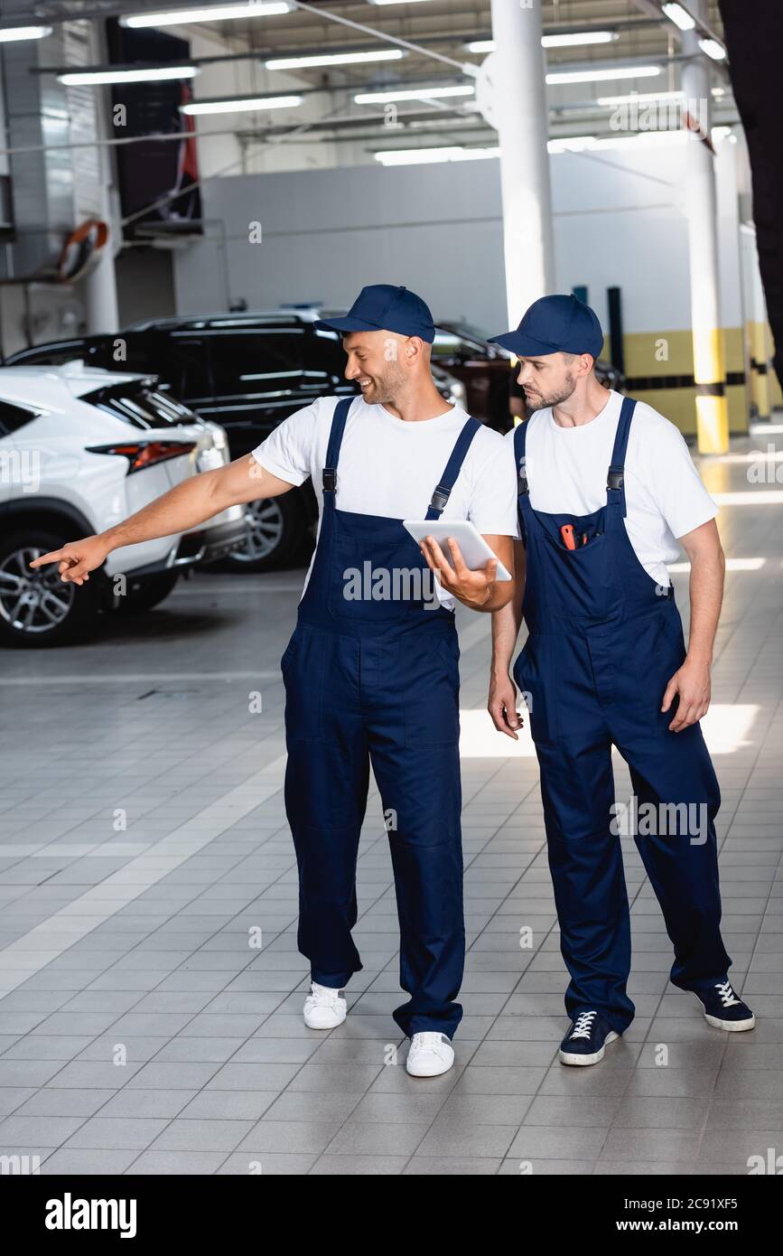 mecánico feliz en uniforme sosteniendo tableta digital y apuntando con el  dedo en el coche cerca de compañero de trabajo en el taller Fotografía de  stock - Alamy