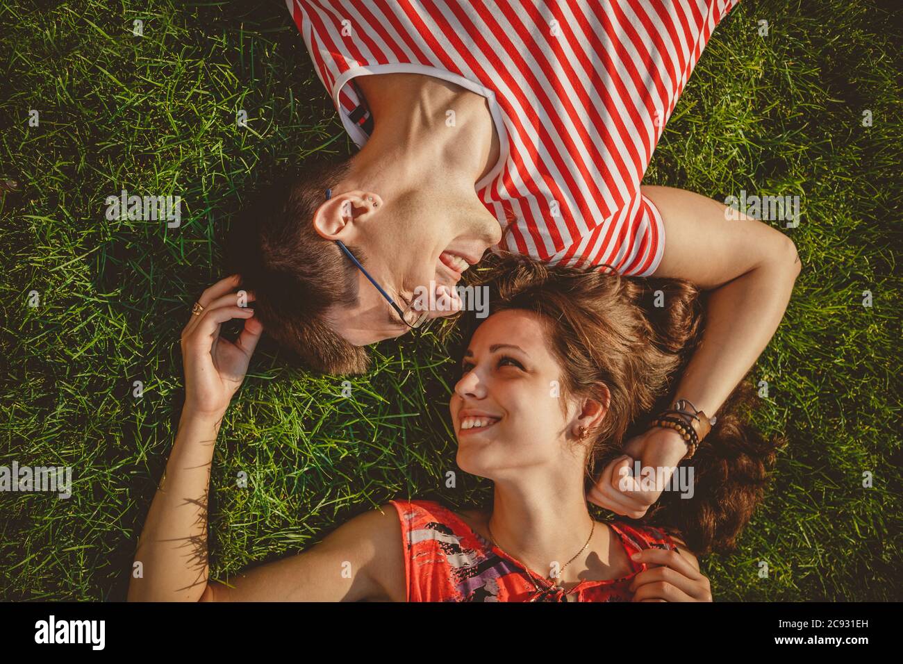 Joven pareja cariñosa acostada juntos cabeza a cabeza en una hierba en verano. Tanto con ropa roja como con las manos. Vista superior Foto de stock