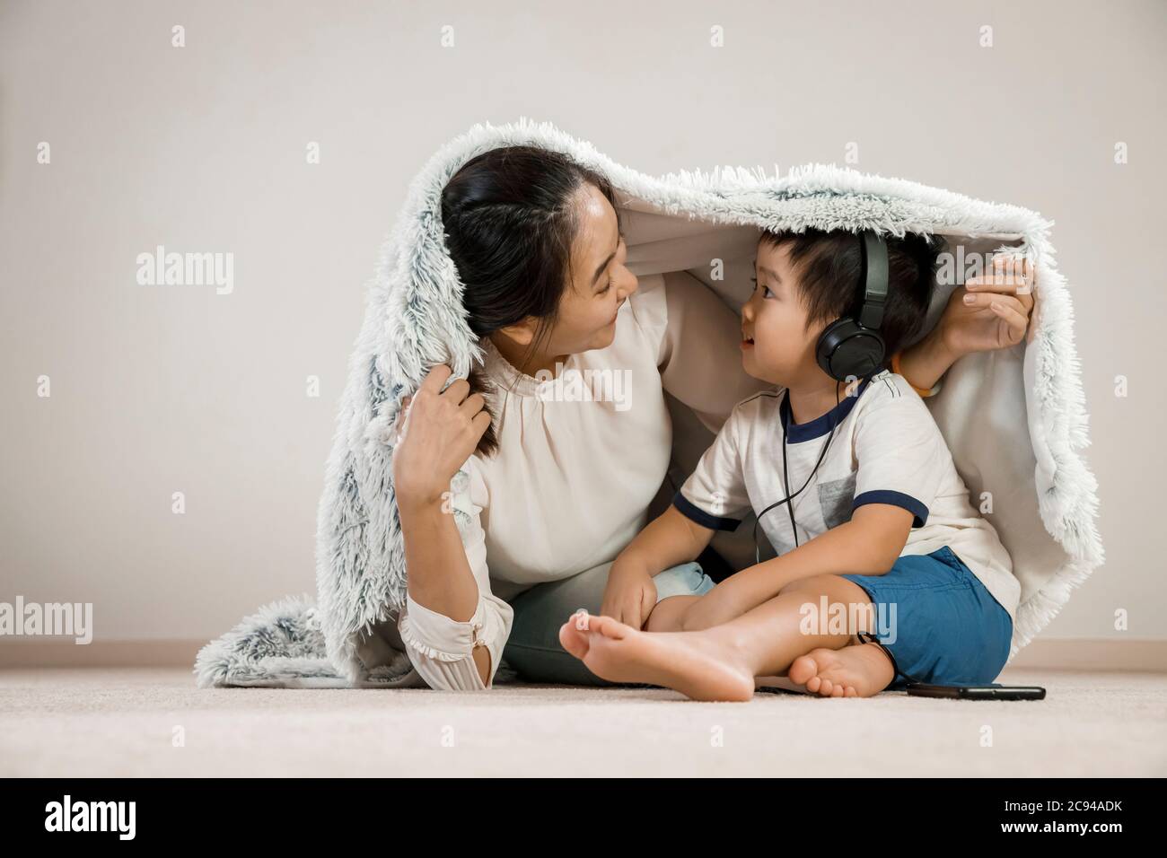 Madre asiática que se esconde bajo una manta con un niño que lleva auriculares. Mamá vietnamita sonriendo jugando ocultar y buscar con su hijo, concepto de tiempo de familia Foto de stock