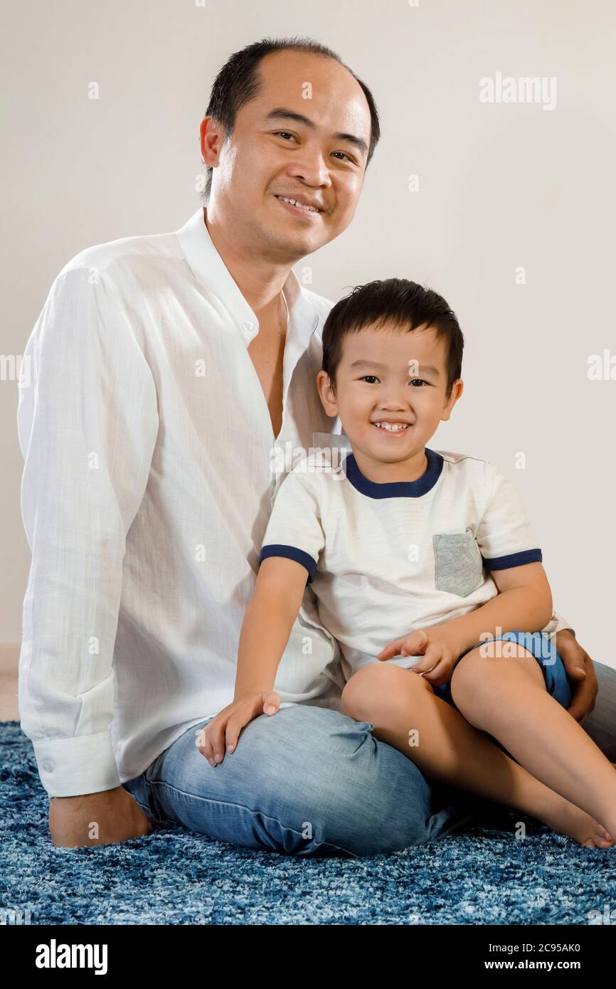 Retrato de padre e hijo asiático feliz y sonriente, hombre vietnamita sentado en el suelo con su hijo en brazos, paternidad o concepto de día de los padres Foto de stock