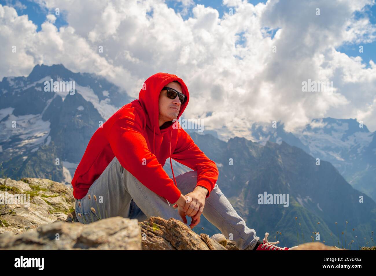 Joven en gafas de sol sentado en la montaña en tiempo soleado. Hombre adulto en hoodie rojo con capucha disfrutando de una hermosa vista en la zona montañosa. Foto de stock