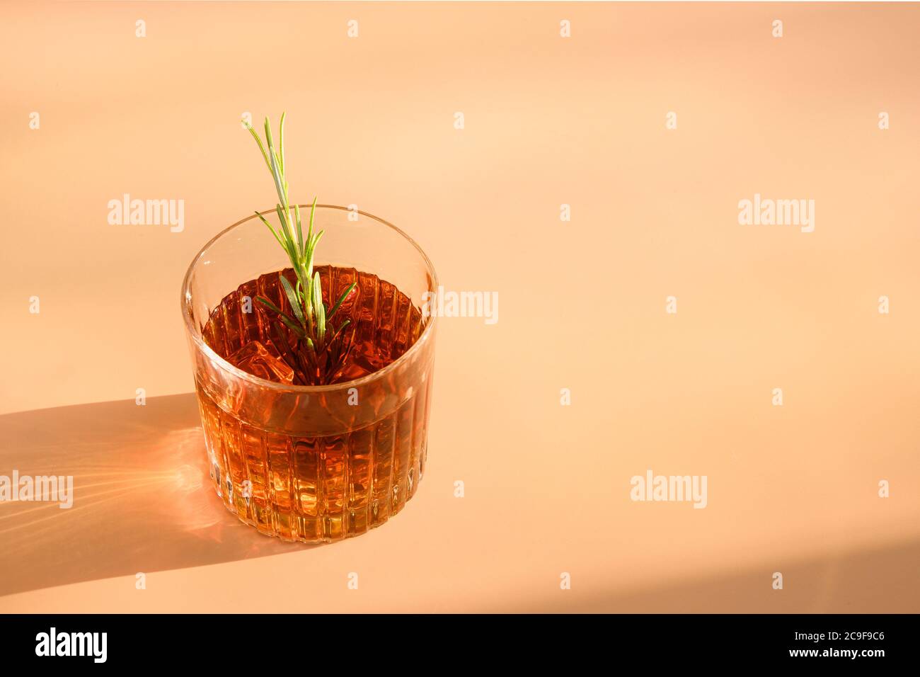 Un vaso de whisky frío adorna el romero sobre fondo beige con sombra soleada. Foto de stock