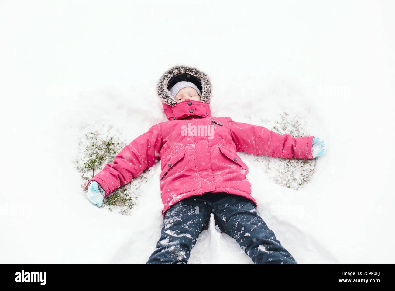 Lindo adorable chica caucásica divertida niña en ropa cálida chaqueta rosa  haciendo ángel de nieve. Niño tumbado en el suelo durante el frío día  nevado de invierno. Niños al aire libre Fotografía