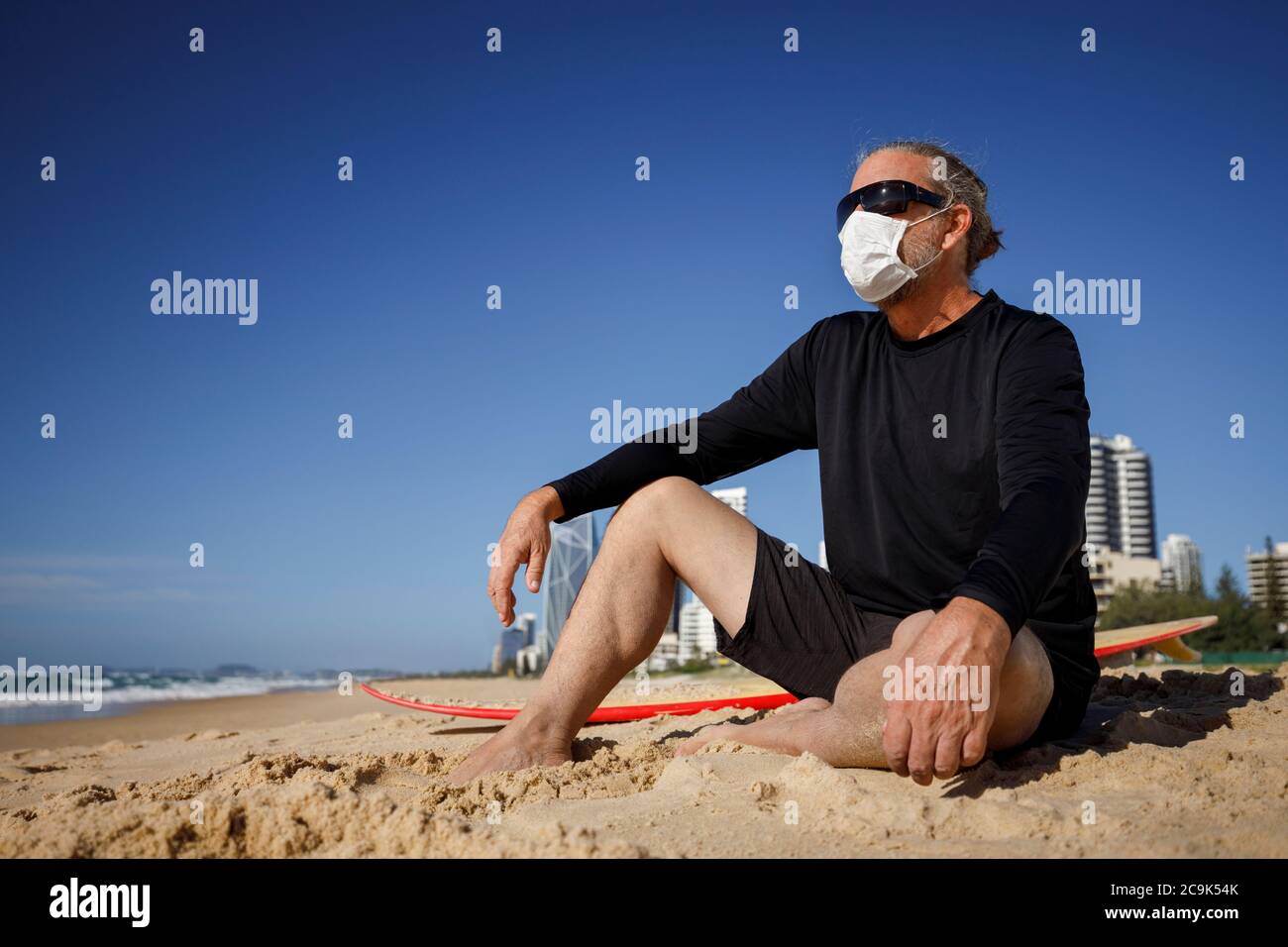 hombre con máscara médica sentado en la playa durante la pandemia de coronavirus covid 19, concepto de distancia social, surfista masculino usando máscara para la protección Foto de stock