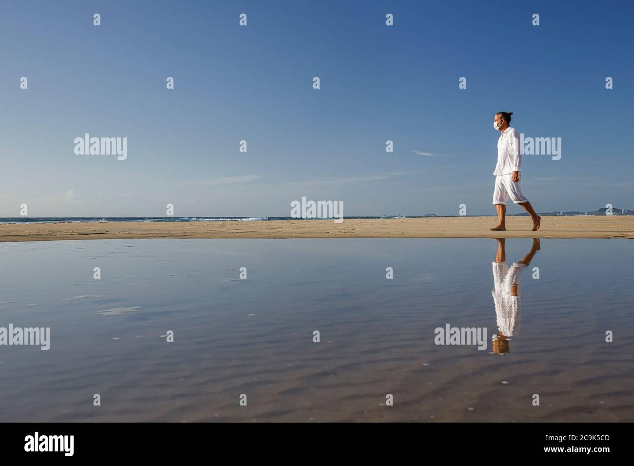 hombre con máscara médica caminando en la playa durante la pandemia del coronavirus covid 19, concepto de distancia social, ejercicio masculino usando máscara para la protección Foto de stock