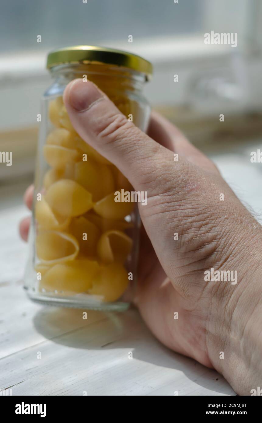 La mano macho sostiene un tarro de vidrio seco de Conchiglie. Porciones de pasta cruda en la mano de un adulto caucásico masculino. Embalaje ecológico para almacenar foo Foto de stock