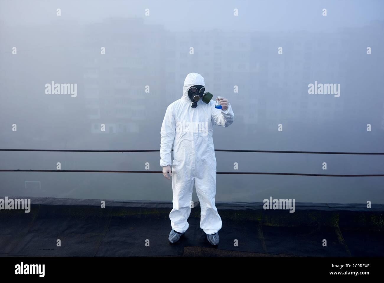 Hombre científico en ropa protectora y máscara de gas, de pie y mirando a un frasco de vidrio con sustancia azul, mala visibilidad a su alrededor, concepto de contaminación del aire Foto de stock