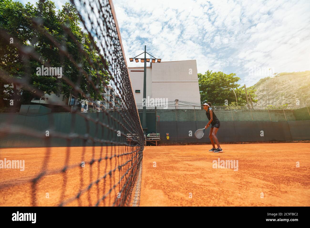 Vista de gran angular de una joven mujer biroquial delgada con una raqueta en las manos de pie sobre una superficie de cancha de arcilla y teniendo una ronda de juego de tenis o un calentamiento Foto de stock