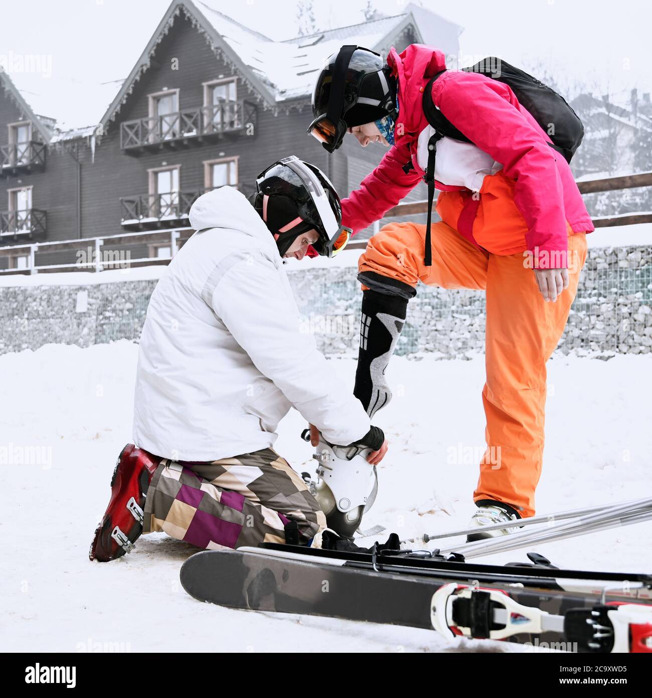Un par de turistas se preparan para esquiar en las montañas en invierno. El  hombre ayuda a su mujer a ponerse botas de esquí y esquís de pie en una  rodilla, amigos