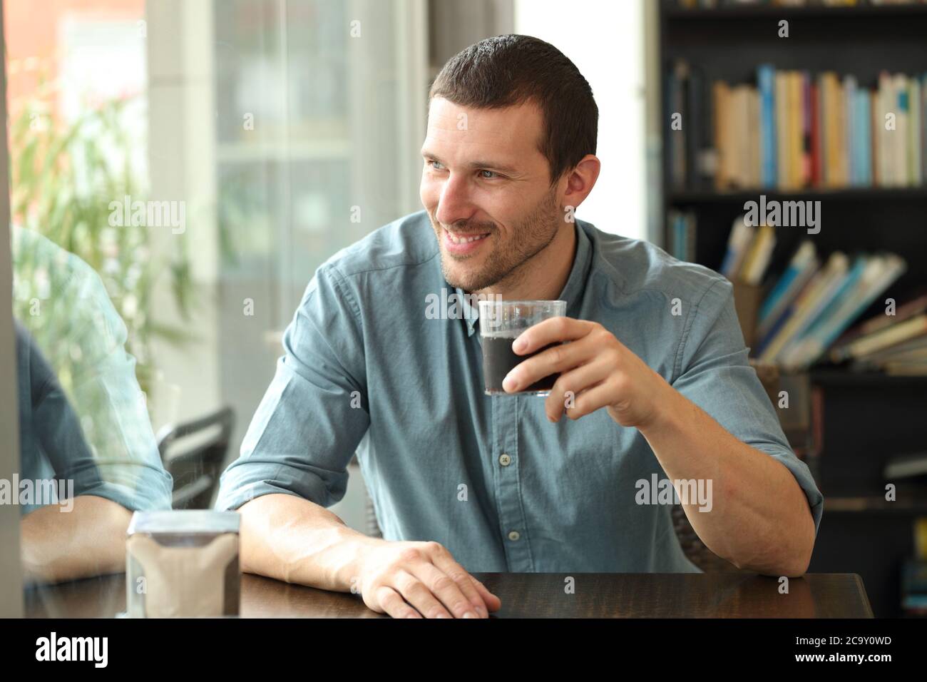Hombre feliz mirando hacia fuera, hay una ventana sentada en una mesa de la cafetería que sostiene un vaso Foto de stock