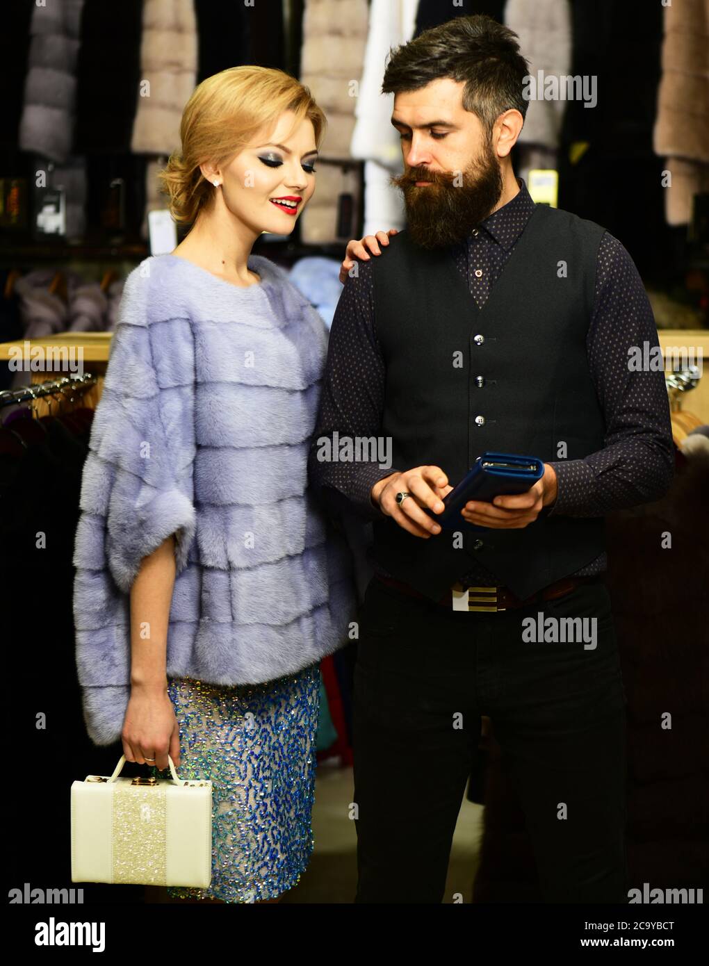 Pareja en el amor intenta abrigos caros. Concepto de lujo y relación. Un chico barba y una mujer compran abrigo peludo. Hombre y niña con caras difíciles sostener la cartera azul