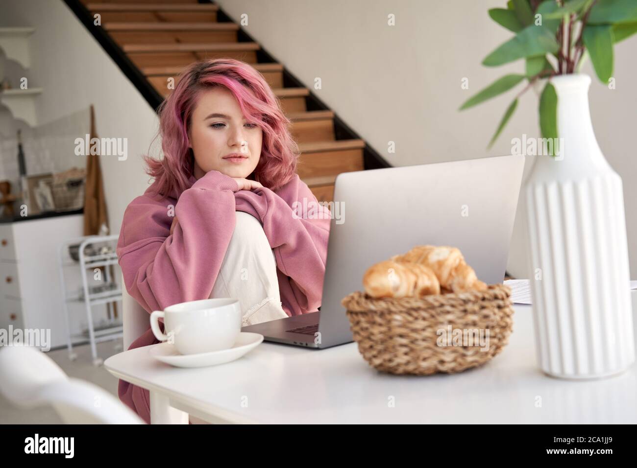Joven hipster chica con cabello rosa viendo video portátil sentarse en la mesa de la cocina. Foto de stock