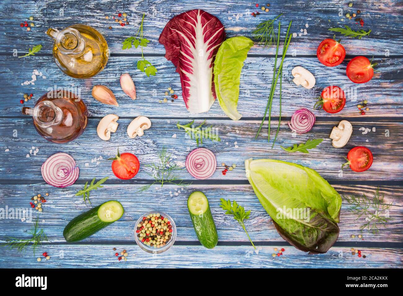 Ingredientes de ensalada de lechuga en una mesa de madera coloreada con tomates, pepino, rucola, granos de sal, cebollino, granos de pimienta, eneldo, perejil, champiñones, rojo Foto de stock