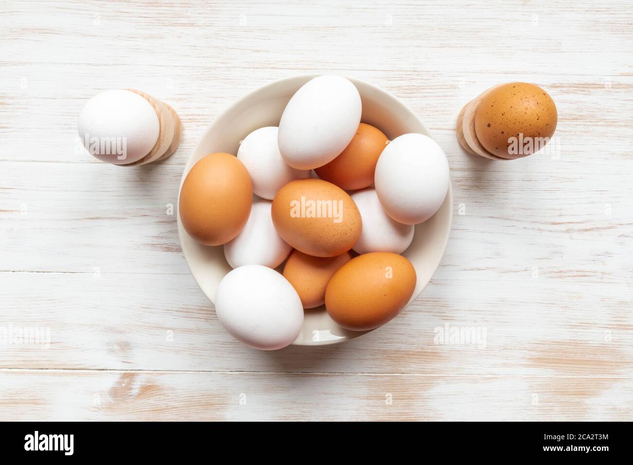 Cuenco con huevos de pollo marrón y blanco sobre mesa de madera. Huevos marrones y blancos en plato y sostenedores sobre fondo de madera. Huevos orgánicos de gama libre. Plana la Foto de stock