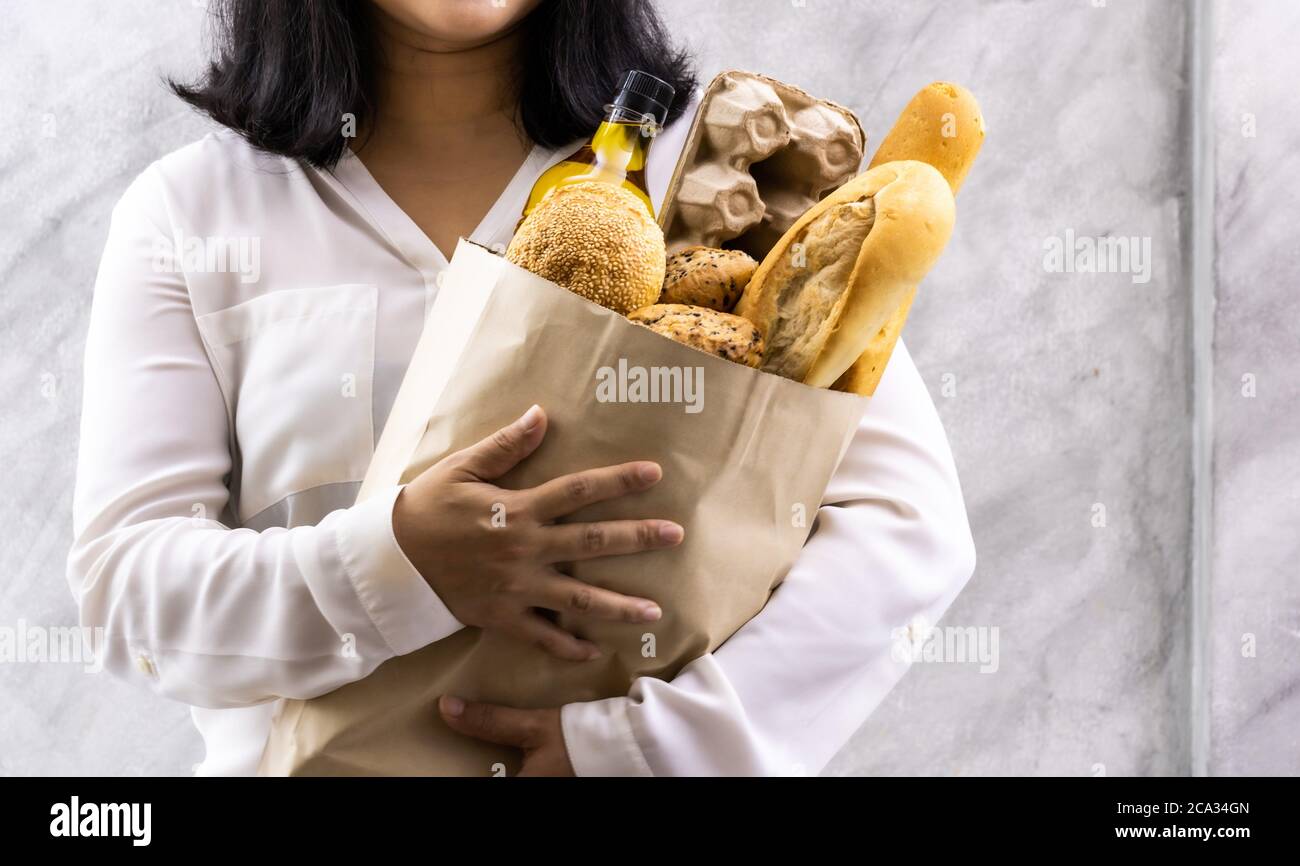 Primer plano de la sonrisa asiática ama de casa sosteniendo pan de variedad en bolsa de papel desechable sobre fondo de loft vintage gris. Panadería y alimentación de bebidas Foto de stock