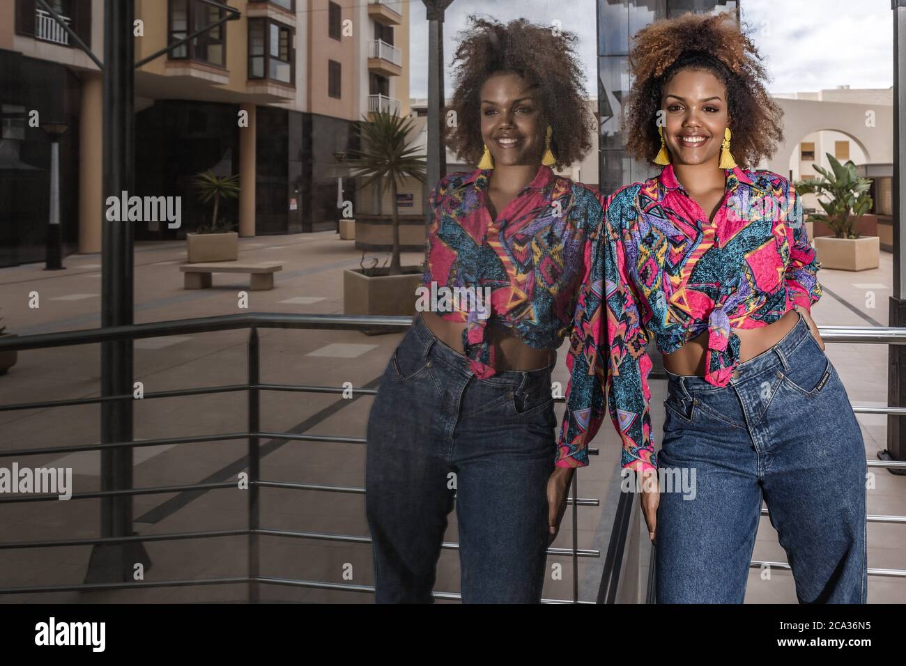 Mujer afroamericana milenaria positiva en elegante camisa colorida con nudo  y jeans de moda que se encuentran cerca de la pared de cristal espejado de  la ciudad moderna Fotografía de stock -