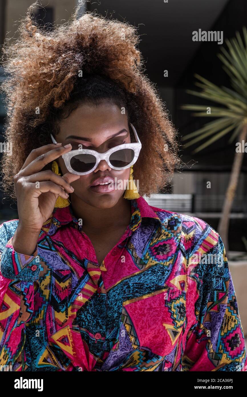 Joven mujer africana americana moderna con un estilo afro peinado en ropa  colorida y con estilo tocando gafas de sol de moda mientras está de pie en  la calle de la ciudad