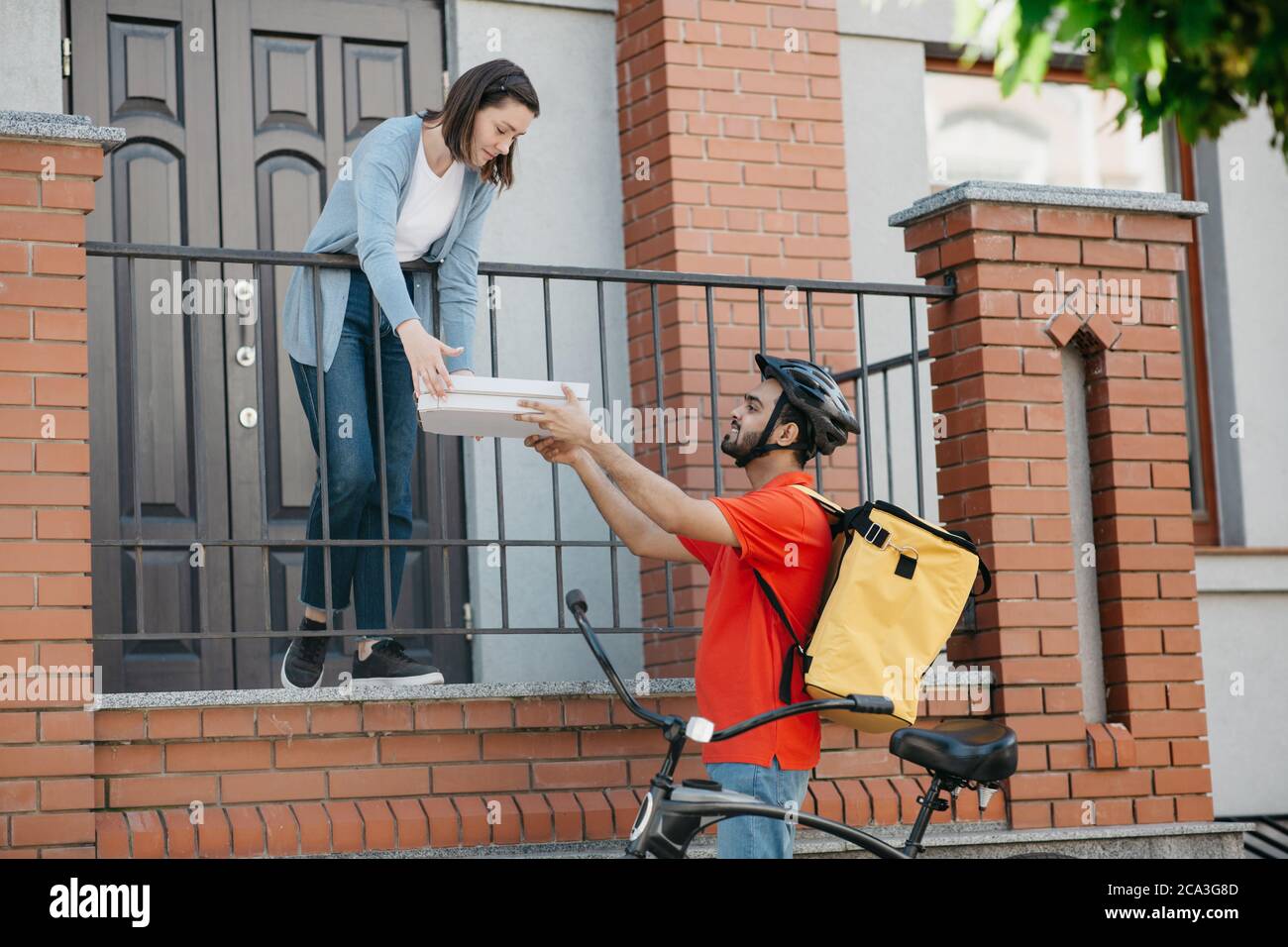Servicio de entrega de comida en la ciudad. La mujer toma la pizza del mensajero de la bicicleta con el casco y la mochila Foto de stock