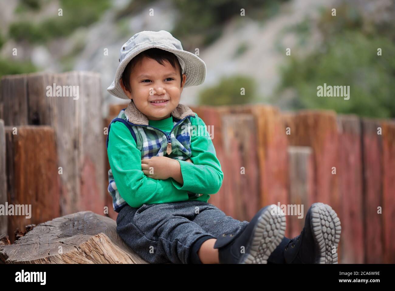 Un niño con los brazos cruzados y sentado, esperando a la cabeza de la trailhead con sus botas de senderismo y sombrero. Foto de stock