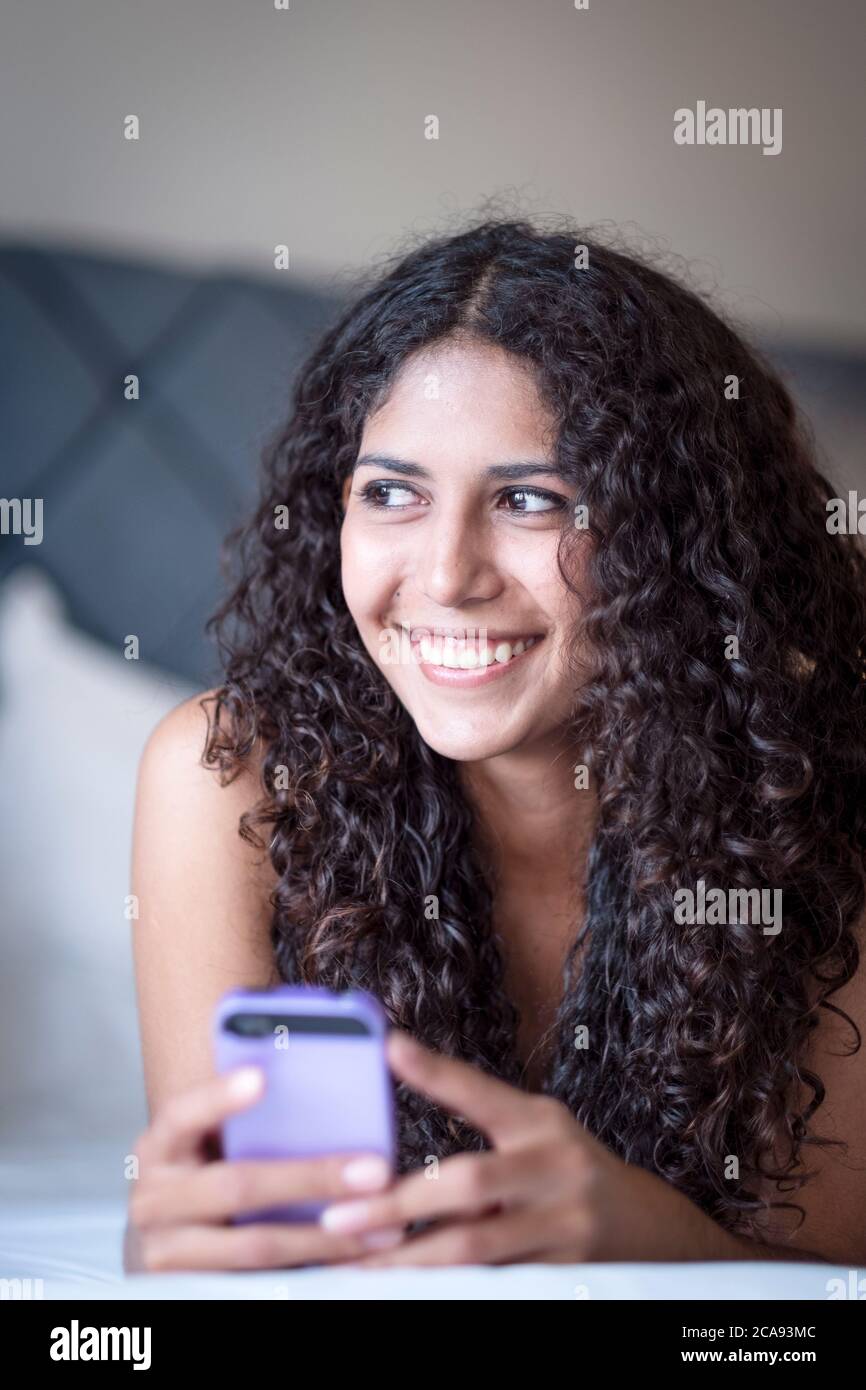 Una mujer latinoamericana acostada en su cama con su teléfono móvil y sonriendo mientras mira fuera de la cámara, Brasil, Sudamérica Foto de stock