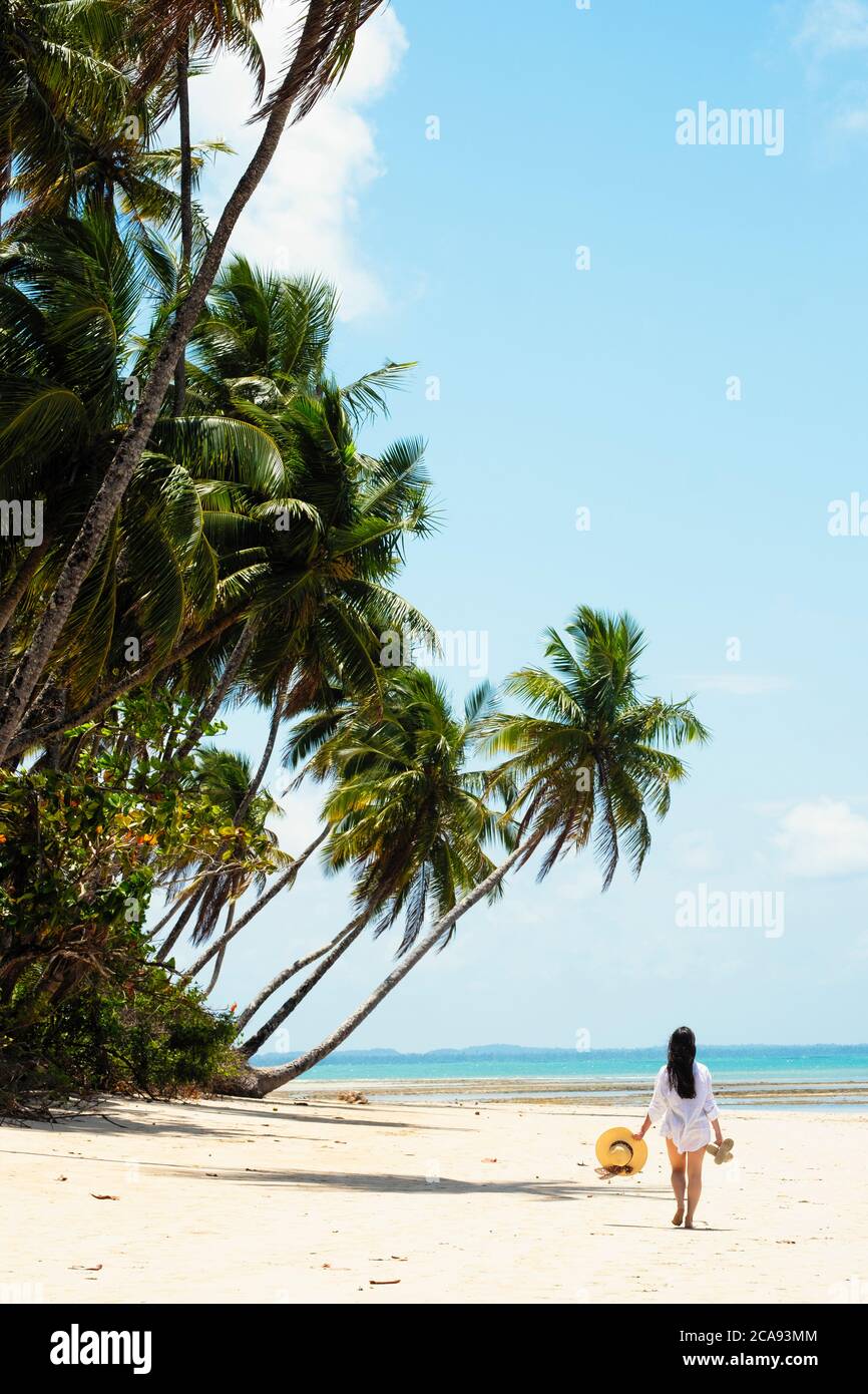 Una mujer joven con pelo marrón con una camisa de playa blanca y sosteniendo un sombrero caminando a lo largo de una playa tropical, Brasil, Sudamérica Foto de stock