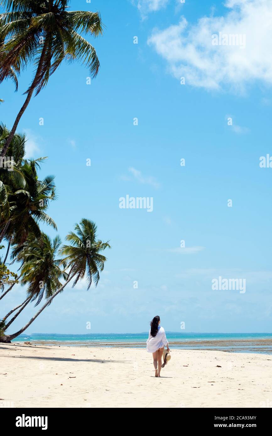 Una mujer joven con pelo marrón con una camisa de playa blanca y sosteniendo un sombrero caminando a lo largo de una playa tropical, Brasil, Sudamérica Foto de stock