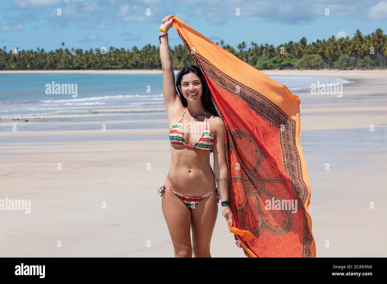 Una hermosa mujer hispana (latinoamericana) en sus veinte años sonriendo, sosteniendo un colorido chal y de pie en una playa desierta, Brasil, Sudamérica Foto de stock