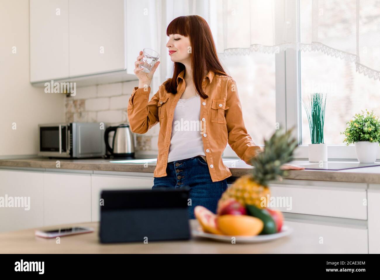 Joven mujer sonriente con ropa casual, bebiendo agua minarl, mientras está de pie en la cocina moderna en casa, comenzando su día activo. Listo para comer Foto de stock