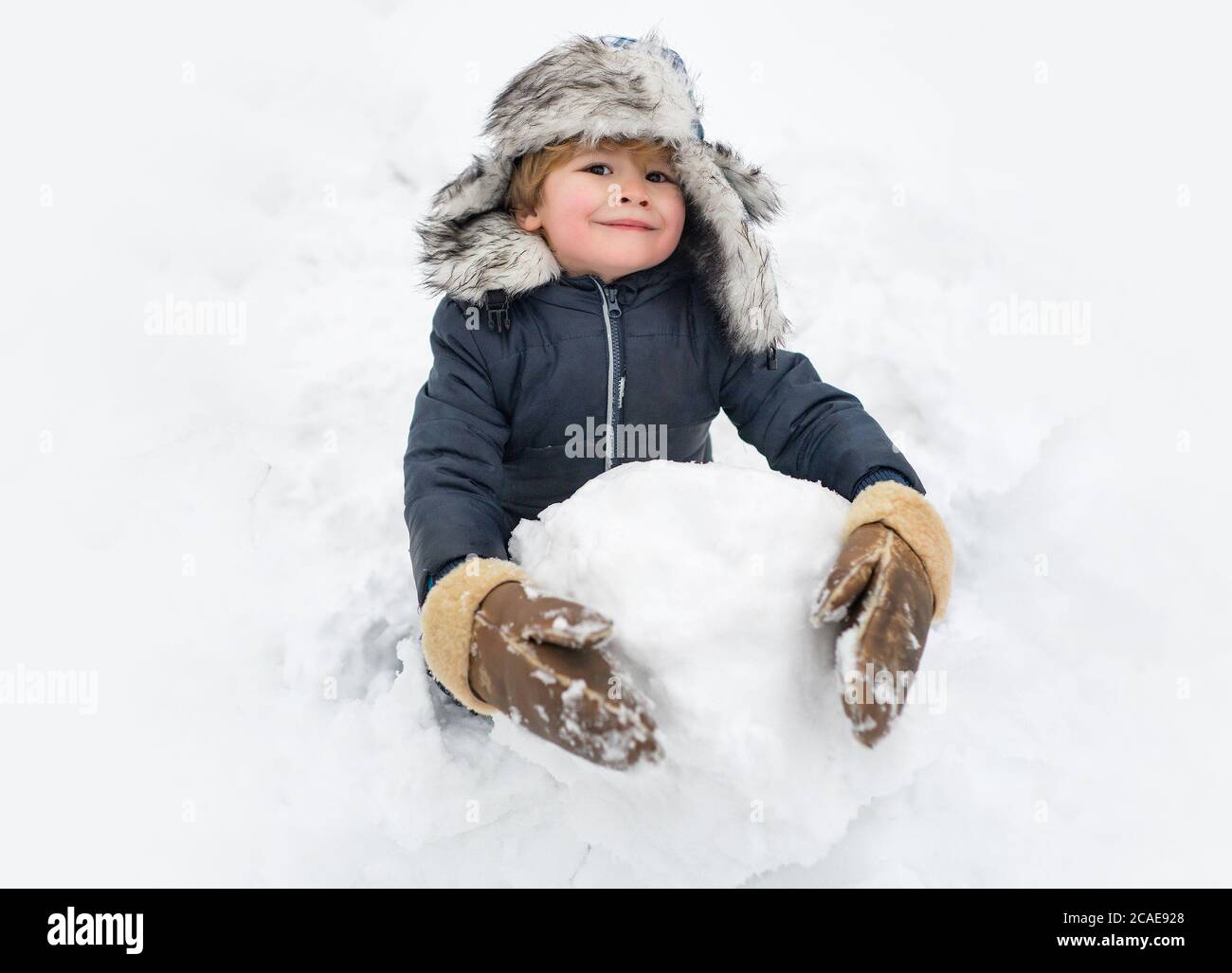 Lindo niño de pie en sombrero de invierno con nariz roja. Niño alegre Diversión y haciendo muñeco de nieve en Winter Park. Ropa de invierno para niños. Foto de stock