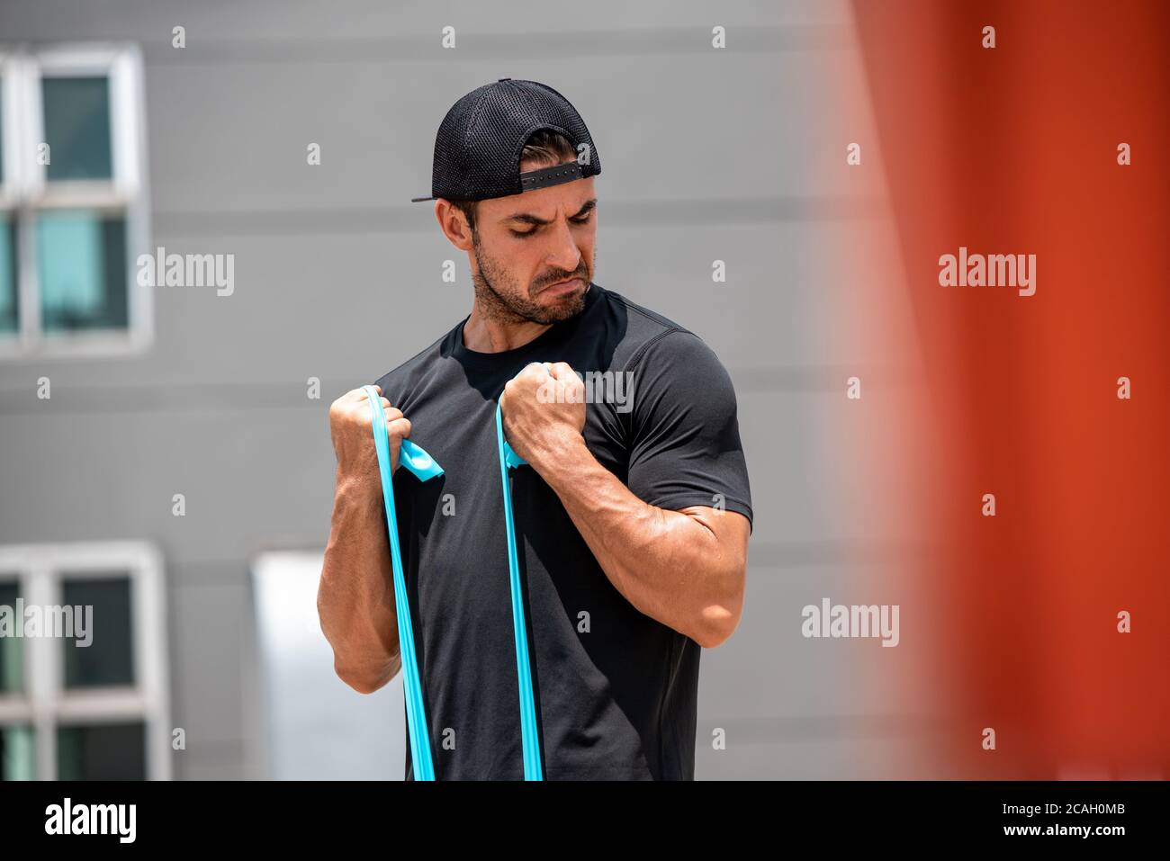 Ajuste muscular hombre deportivo latino haciendo ejercicio de rizo bíceps con banda de resistencia al aire libre en casa bajo la luz del sol Foto de stock