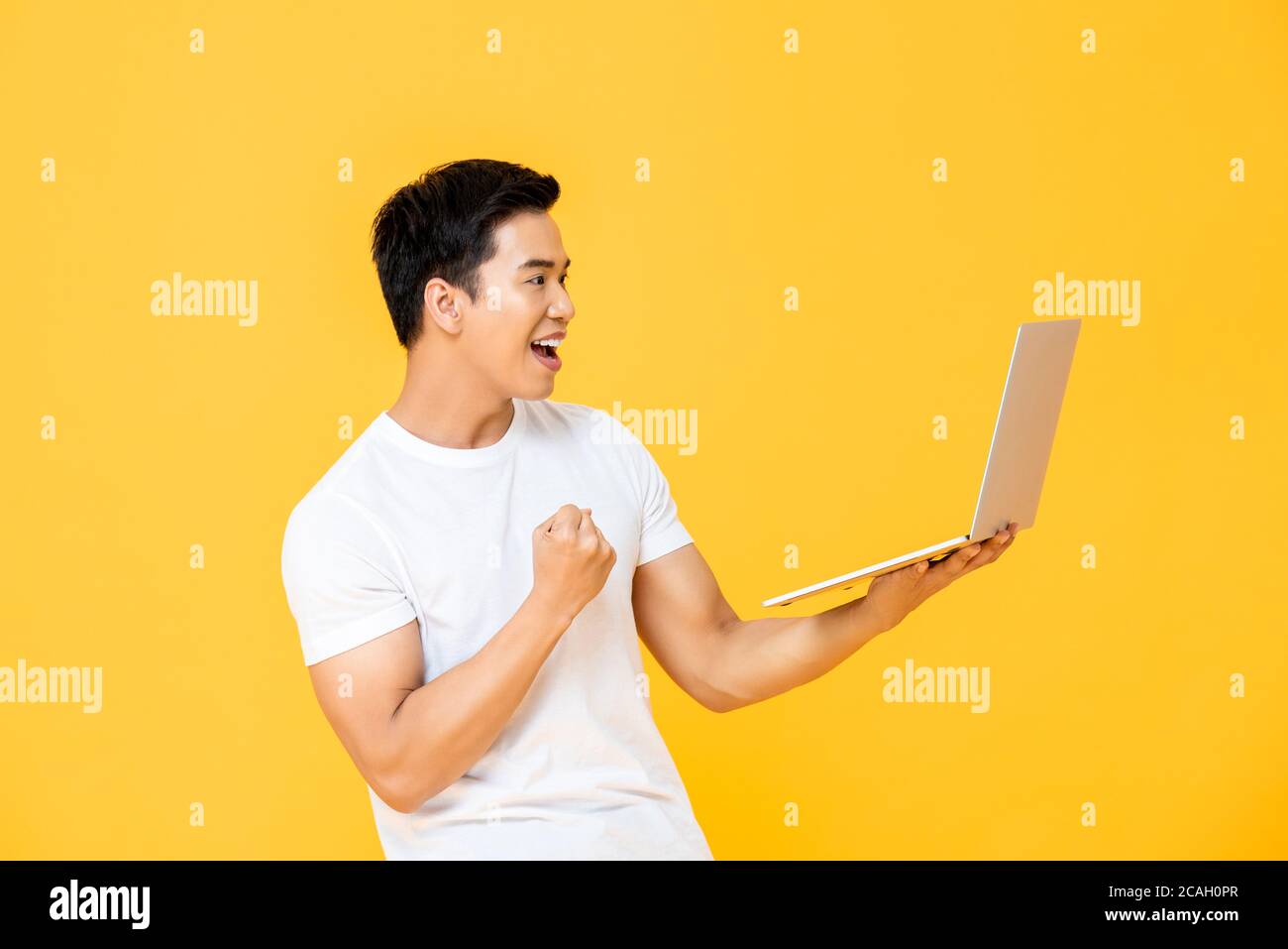 Un joven asiático emocionado mirando la computadora portátil y criando su puño haciendo sí gesto aislado sobre fondo amarillo Foto de stock