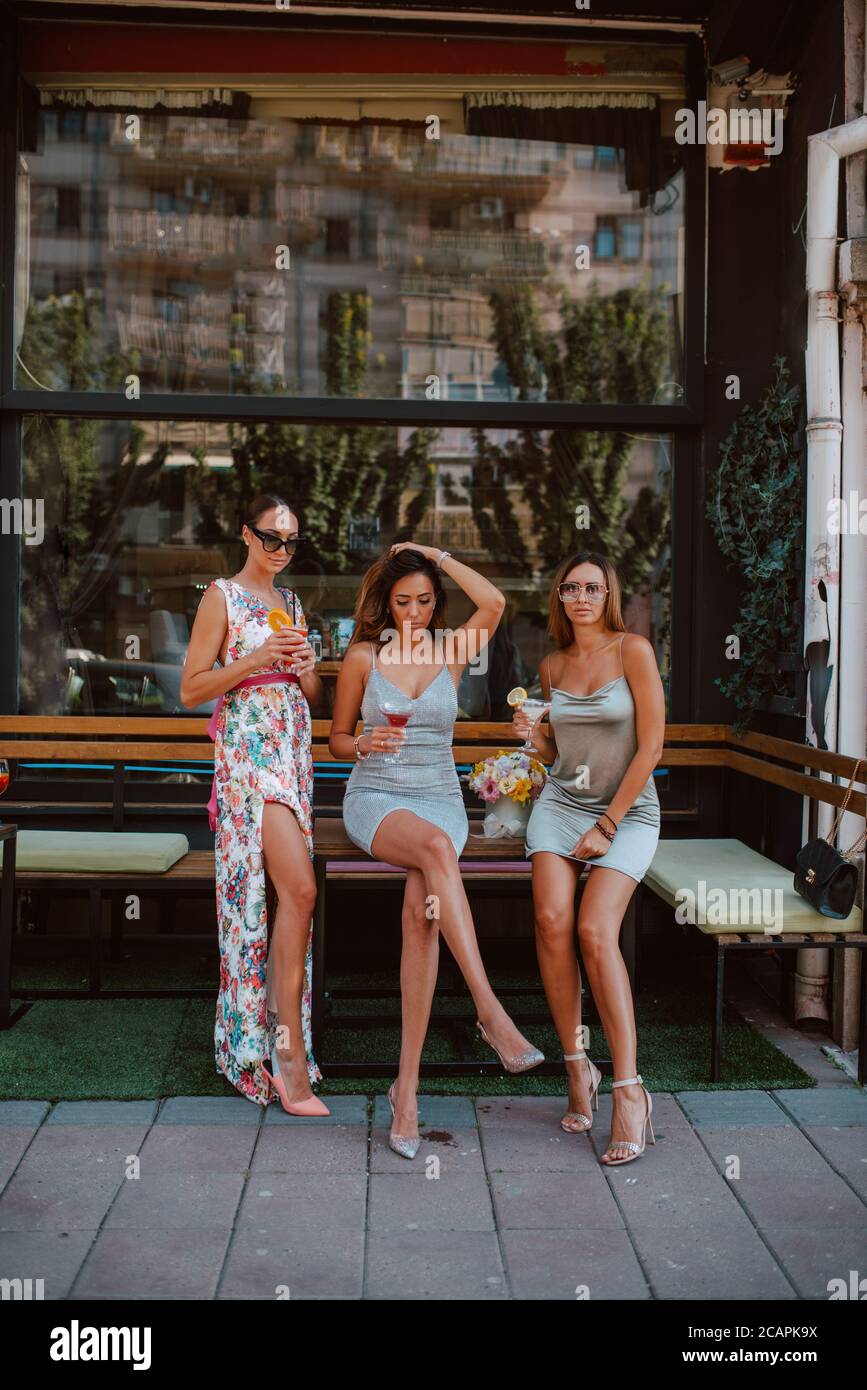 La longitud de tres mujeres jóvenes caucásicas hermosas en vestidos cortos de verano están bebiendo cócteles en un jardín café. Chicas de fiesta Fotografía de stock - Alamy