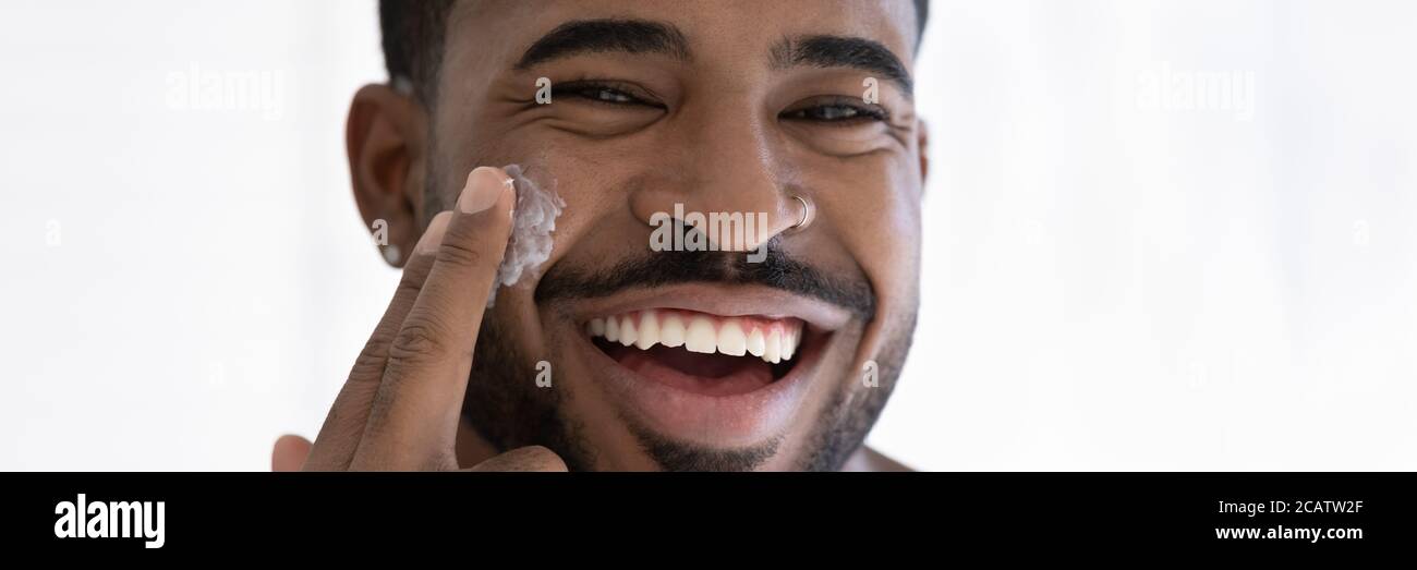 De cerca un hombre afroamericano demasiado alegre aplicando crema facial hidratante Foto de stock