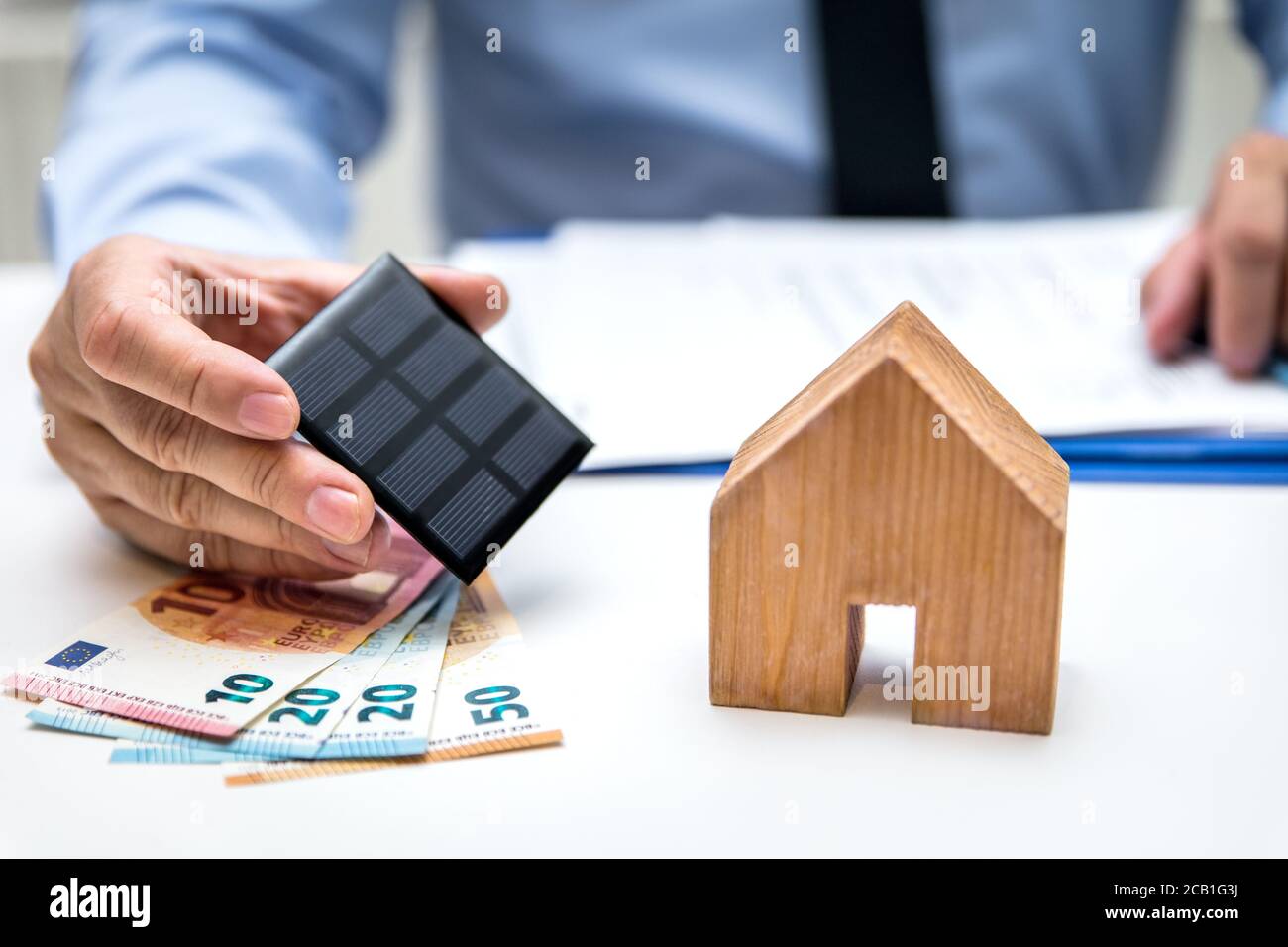 Hombre con billetes en euros y un panel solar en su mano, casa al lado, energía verde y electricidad Foto de stock