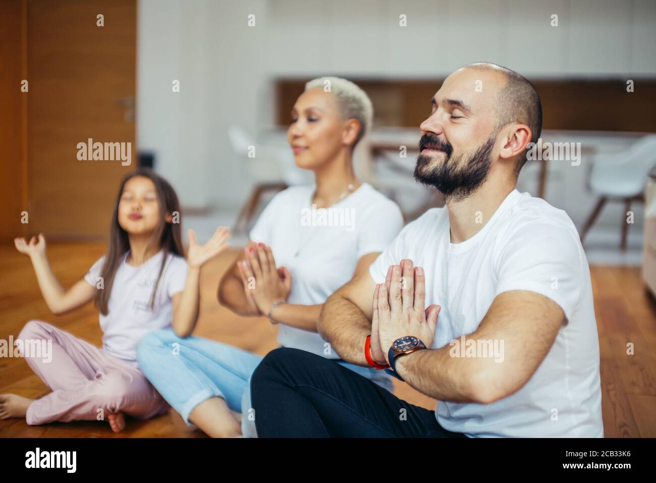 Padres jóvenes caucásicos con niño meditando en casa aislados en la habitación, sentados en el suelo. Familia, deporte, yoga concepto. Colores blancos Foto de stock