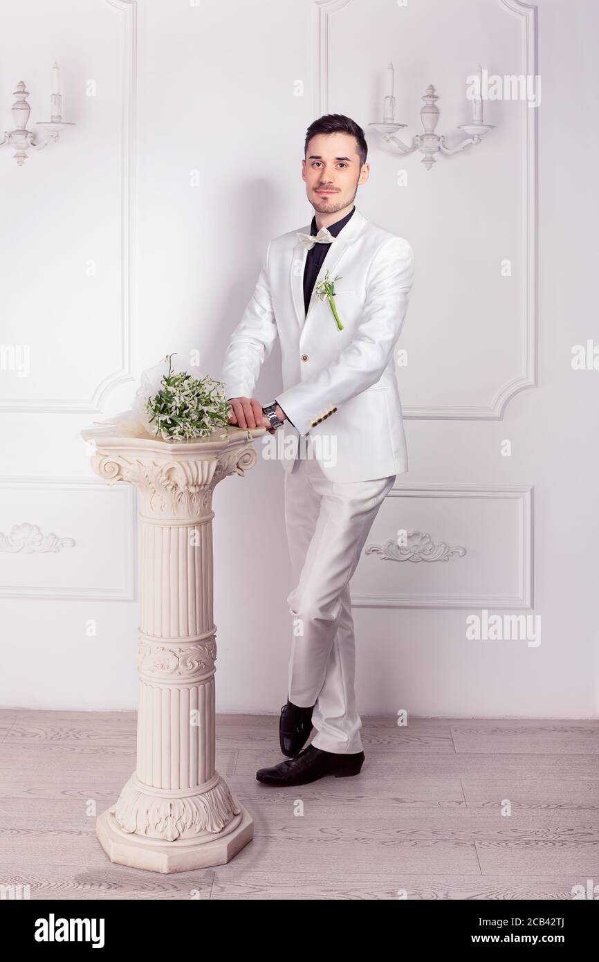 Moderno novio en traje blanco y zapatos negros de pie con ramo de novia en  una pequeña columna cerca de mirar la cámara Fotografía de stock - Alamy