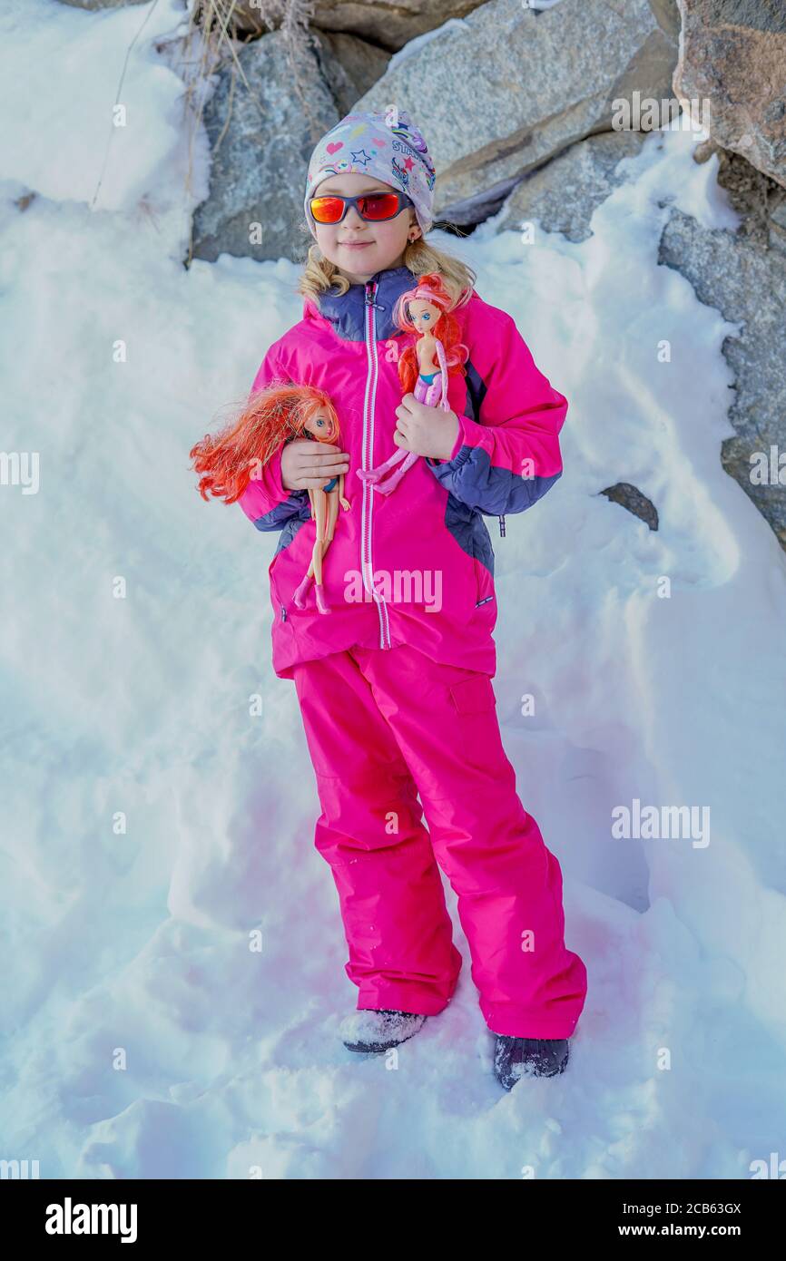 L'Alpe D'Huez, Francia 30.12.2019 Retrato de invierno al aire libre de niña pequeña y linda con gafas de sol y ropa de esquí. Chica con muñecas. Ropa elegante para niños Fotografía
