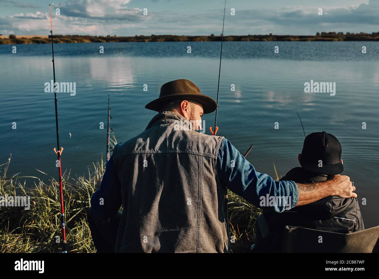 Las espaldas del padre y el hijo caucásicos se sientan cerca del lago en sillas de pesca. Están felices de pasar tiempo juntos. Cálido día soleado. Foto de stock