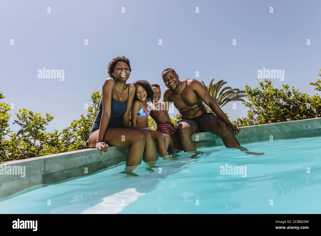 Feliz familia joven sentada en el borde de la piscina. Familia joven disfrutando de sus vacaciones de verano. Foto de stock