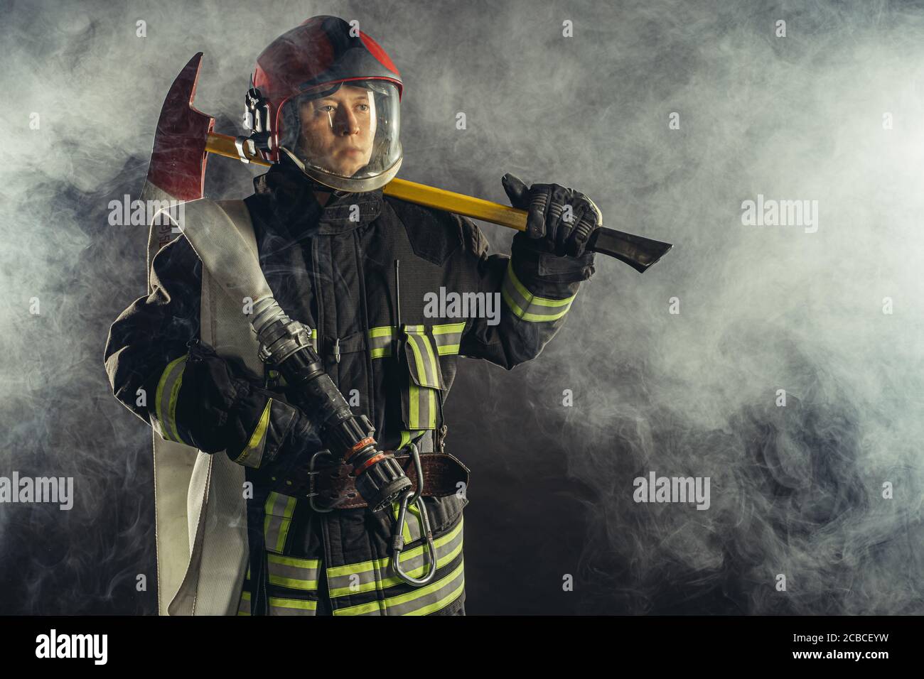 joven bombero caucásico sosteniendo un martillo, arriesgando su vida para salvar a la gente del fuego, usando uniforme protector Foto de stock
