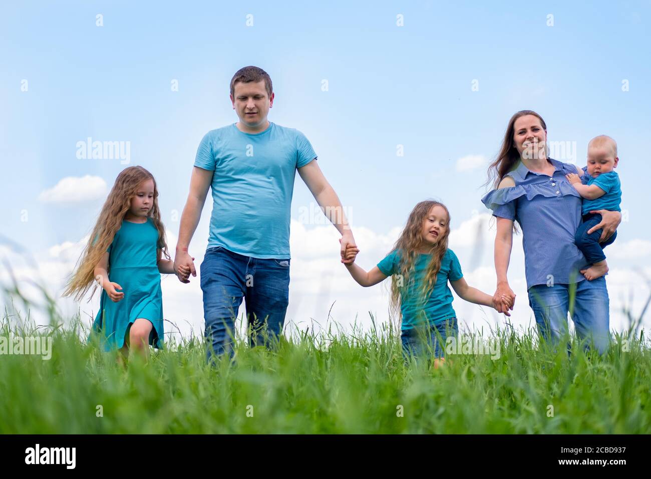 Papa Mama Y Tres Ninos Caminan Sobre Hierba Verde Contra El Cielo Azul Felices Padres Caucasicos Dos Hijas Y Un Hijo Pequeno Que Se Agarran De Las Manos Fotografia De Stock